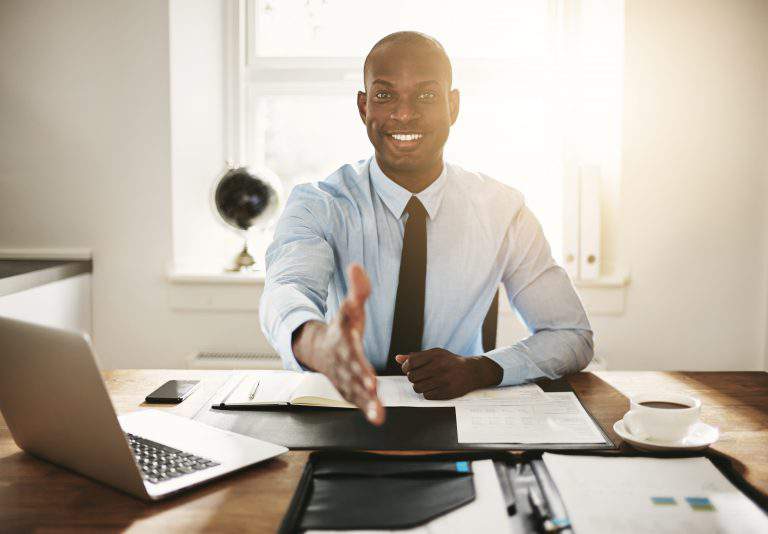 Smiling young African executive extending his arm in a handshake while sitting at his desk in an office